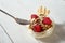 Bowl with dessert with whipped cream, raspberries and nuts with spoon on white wooden table.