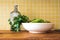 A bowl of green vegetables  and a glass bottle against a yellow tiled wall in a kitchen