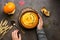 A bowl of hot pumpkin cream soup with sour cream and seeds in woman hands in a woolen sweater on a dark rustic background. Autumn