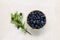 Bowl of tasty fresh bilberries and green leaves on white table, flat lay