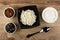 Bowls with raisin and prune, bowl with grainy cottage cheese, bowl with sour cream, spoon on wooden table. Top view