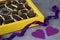 Box of chocolates, two purple decorative hearts and purple ribbon on gray background.