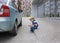 Boy of 4-5 years old in medical mask sits alone on the sidewalk near a car and a building