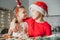 Boy 8 years old in red Santa hat hugs dark-haired girl 3 years old. Siblings in Christmas kitchen look at each other