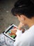 Boy with black short hairs wearing white casual pyjamas sitting outdoor concentration on playing colourful interface game on a han