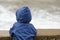 Boy in blue jacket with hood stands with his back against the pier against the background of sea waves