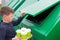 A boy in a blue jumper throws disposable glasses into a green dumpster