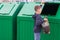 A boy in a blue jumper throws a full bag of garbage into a green container