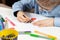 Boy in a blue shirt and glasses sits at a white desk and draws with crayons on a white piece of paper. In the foreground colorfu