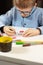 Boy in a blue shirt and glasses sits at a white desk and draws with crayons on a white piece of paper. In the foreground colorfu