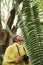 Boy With Camera Looking At Large Fern In Forest