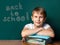 A boy of Caucasian appearance on the background of the school board. Next to him are a stack of notebooks and textbooks. Looking