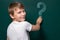 A boy of Caucasian appearance writes a question mark on the chalkboard. Looking into the camera. Smile. Easy solution to the