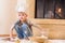 A boy in chef`s hats near the fireplace sitting on the kitchen floor soiled with flour, playing with food, making mess