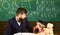Boy, child in graduate cap looks at scribbles on chalkboard while teacher explains. Teacher with beard, father teaches