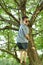 Boy in dark sunglasses climbs tall tree, clings to thick branches, concept of summer relaxation, dangerous outdoor activities,