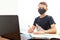 A boy in a disposable mask at home in front of a computer does his homework. Distance learning in quarantine. Textbooks are on the