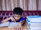 Boy doing homework and reading on a wooden table with a pile of books beside The background is a red sofa and cream curtains