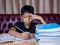 Boy doing homework and reading on a wooden table with a pile of books beside The background is a red sofa and cream curtains