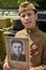 Boy dressed in Soviet military uniform during the second world war posing near army tank and holds portrait his grandfather who