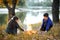 Boy and girl sitting on the river Bank, make a fire, autumn forest at sunset, beautiful nature and reflection of trees in the wate