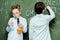Boy and girl in white coats standing with reagents in flasks and drawing chemical formulas