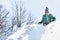 The boy in hat, scarf and green jacket slides down from a snow slide on back. Winter day