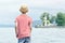 Boy in hat standing on the beach. Small lighthouse in the distance. Back view