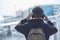 A boy in headphones with a backpack stands near the window at the airport and looks at the plane.