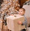boy, holding a huge box with a gift for the new year, sits in a light chair, against the backdrop of a Christmas tree