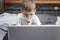 A boy in a medical mask sits in front of a laptop at home in bed. Distance learning