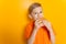 Boy in an orange T-shirt holds a hamburger in both hands and greedily bites it on a yellow background