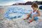 A boy is played with seashells and sand while sitting on a beach strewn with jellyfish near the sea