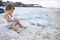 A boy is played with seashells and sand while sitting on a beach strewn with jellyfish near the sea