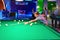 The boy plays billiards and hits the ball with a cue, driving him into a pocket on a green table