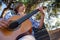 Boy plays the guitar in the park sitting on a bench.  Child plays a musical instrument, in nature.  Guitarist plays on guitar