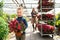 Boy with a potted plant and his mom with a cart with flowers in a greenhouse from behind.