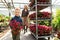 Boy with a potted plant and his mom with a cart with flowers in a greenhouse from behind.