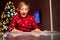 A boy in a red Christmas sweater on the background of a Christmas tree draws in front of a lamp at a white table