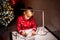 A boy in a red Christmas sweater on the background of a Christmas tree paints a drawing with felt-tip pens