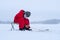 A boy in red jumpsuit on a frozen snowy lake. Fishing rod in the hands of a boy. Looks into the hole.