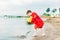 Boy in a red raincoat pours water out of yellow rubber boot into the lake. Child playing with water at pond. Kid having