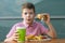 A boy in a red shirt eats a sandwich and drinks a drink through a straw from a glass at a table in a cafe