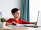 A boy in red shirt a hand on computer`s mouse focusing on study writing on paper, homeschool concept.