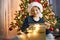 A boy in a santa hat opens a gift box next to a garlanded Christmas tree. Gifts for children. Dreams Come True