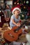 Boy in Santa hat is playing in front of a decorated Christmas tree with guitar