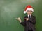 Boy in Santa red hat points on empty green chalkboard