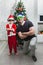 A boy in a Santa suit and his dad in funny sunglasses are on one knee near the Christmas tree with their arms crossed on their