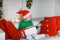 A boy in a santaclaus cap and a green sweater sits on a sofa and holds a white box with a gift