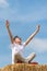 Boy screams with his hands up sitting on the haystack. Portrait of teenager on sheaf of hay. Blue summer sky. Vertical frame